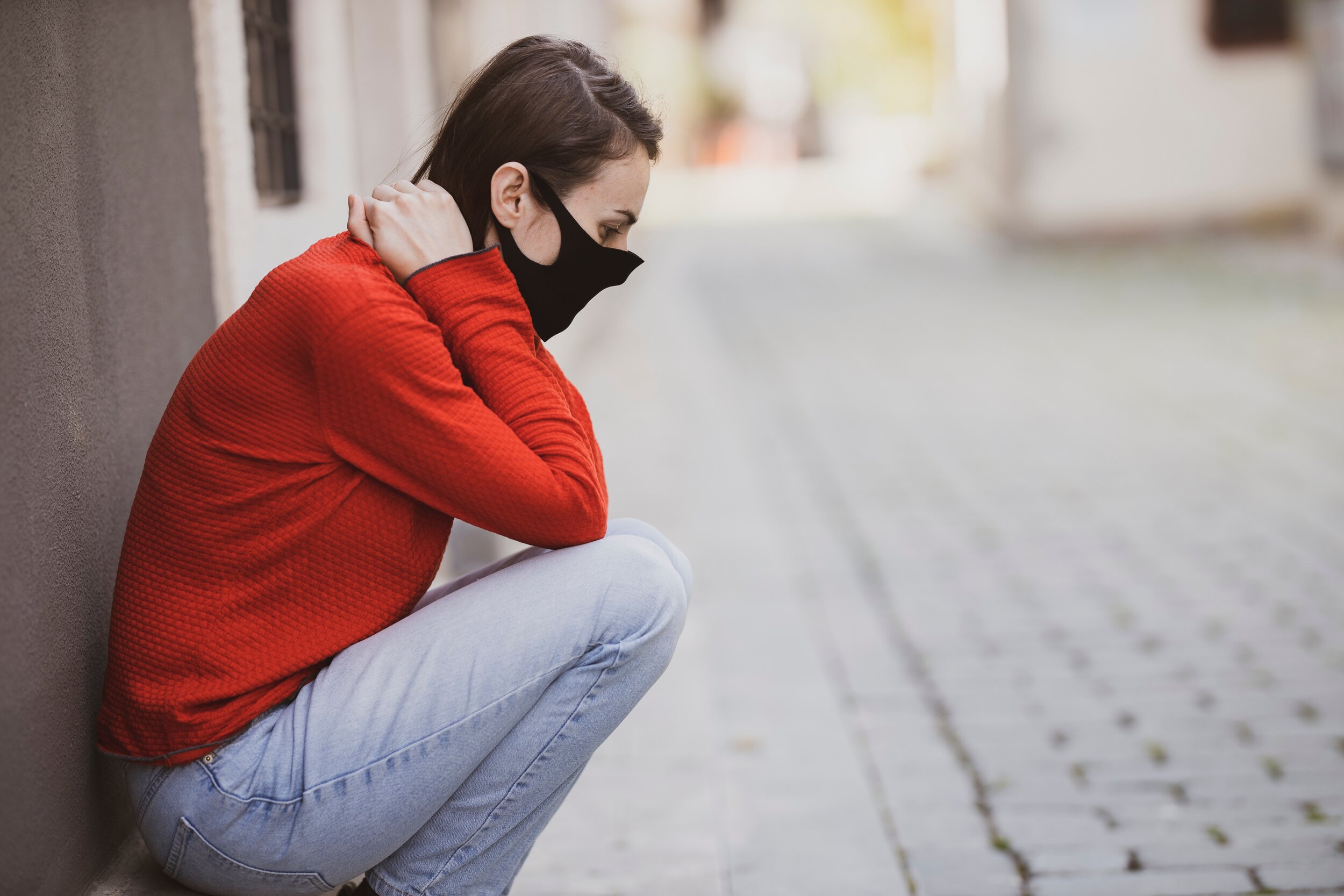 Woman in black mask and red sweater crouching on cobblestone street anxiously