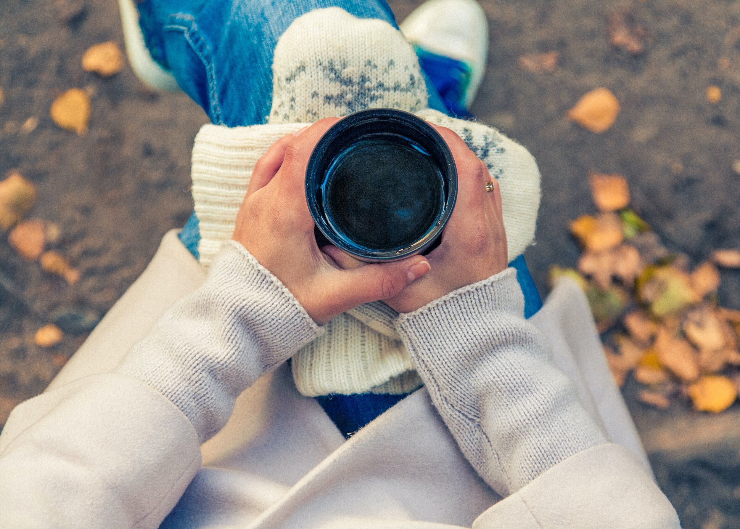 Person in cream sweater holding hot drink surrounded by autumn leaves