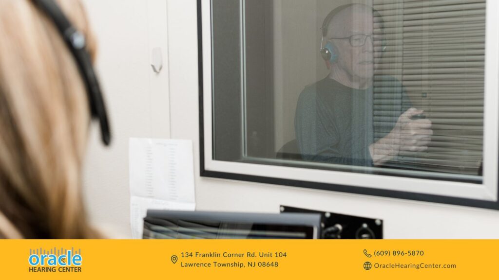 Audiologist conducting a hearing test while an elderly man sits in a sound booth wearing headphones.